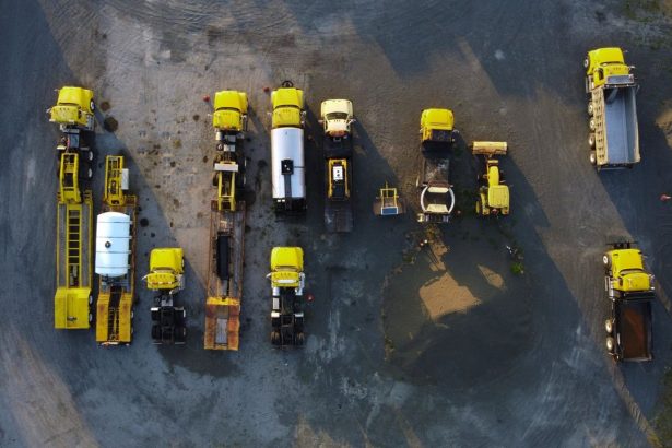 Vista aérea de caminhões e máquinas de construção amarelos estacionados em fileiras sobre um terreno de cascalho.