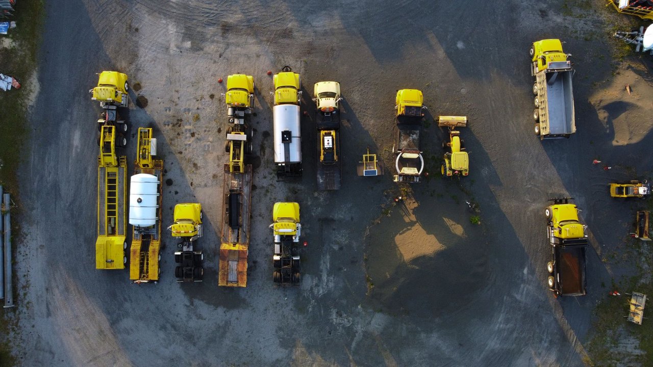 Vista aérea de caminhões e máquinas de construção amarelos estacionados em fileiras sobre um terreno de cascalho.
