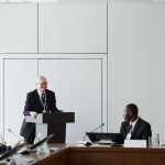 Grupo de profissionais participa de uma reunião formal em uma sala de conferências, com um participante falando ao púlpito.