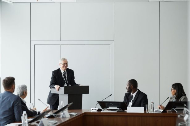 Grupo de profissionais participa de uma reunião formal em uma sala de conferências, com um participante falando ao púlpito.